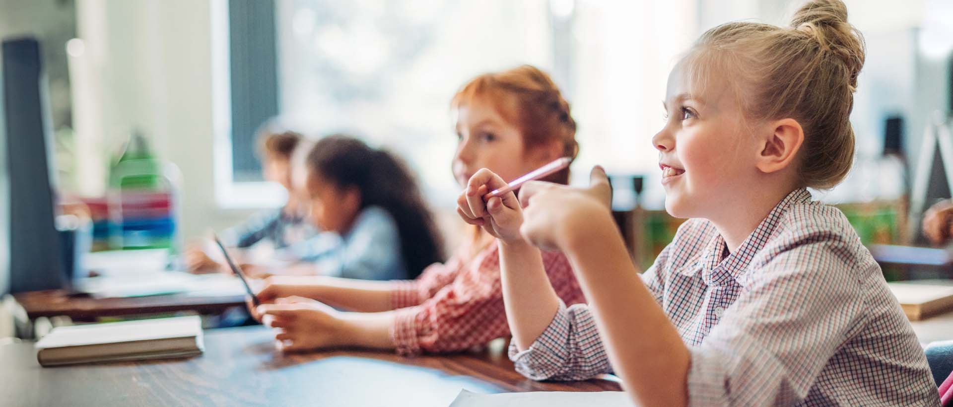 Bildet viser en ung jente i en klassemiljø, som smilende lytter og holder en blyant. Dette illustrerer kanskje et interaktivt og samarbeidsbasert læringsmiljø, muligens knyttet til bruk av programvaren Flyt Skole og dens spesifikasjoner for samarbeid mellom elever.